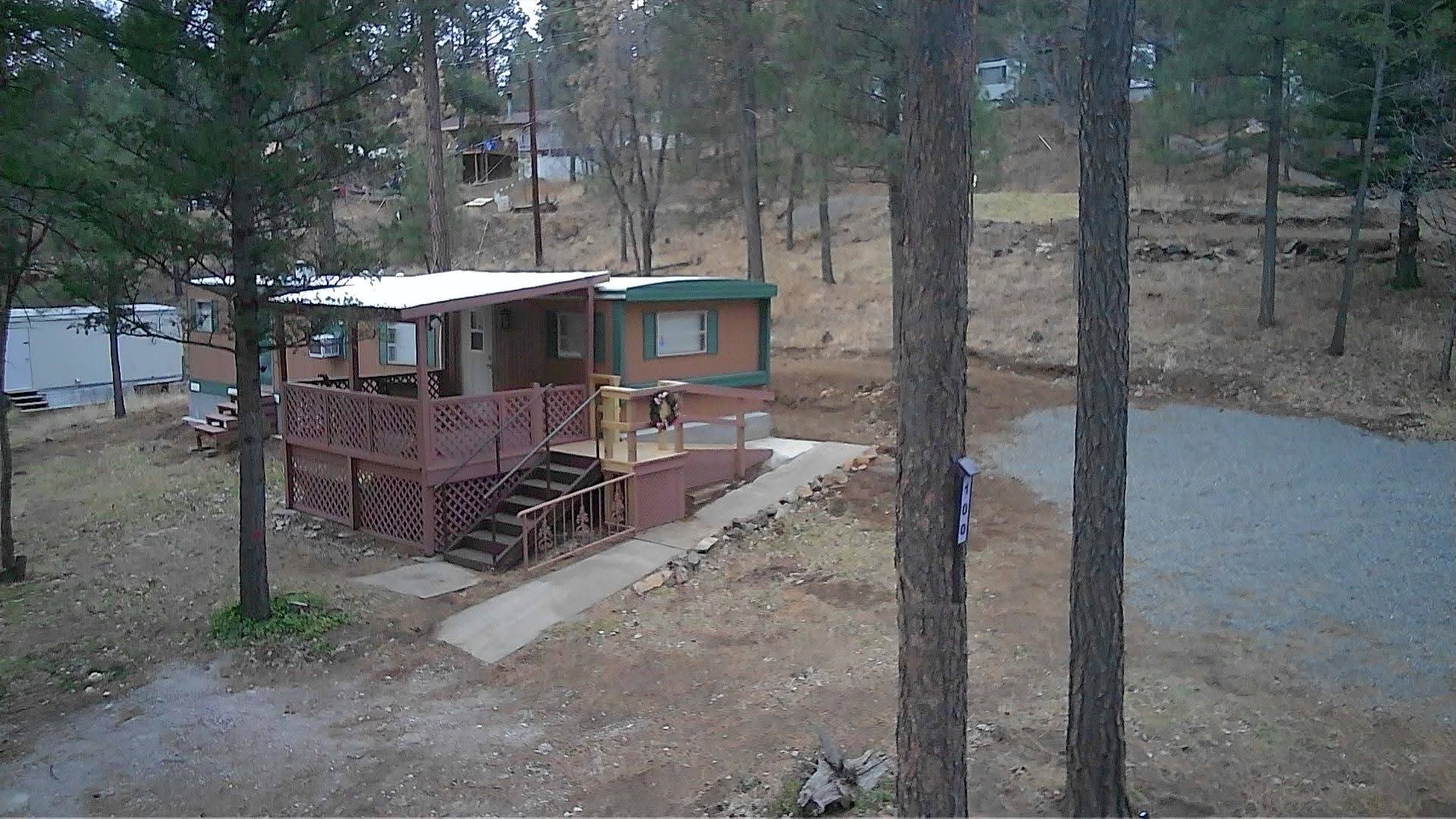 Scenic aerial perspective of the cabin nestled among tall pine trees, surrounded by forested landscape with distant mountains and clear sky, showcasing the peaceful natural setting